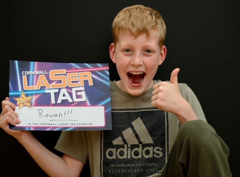 Young boy sat smiling and doing a thumbs up with his laser tag champion certificate