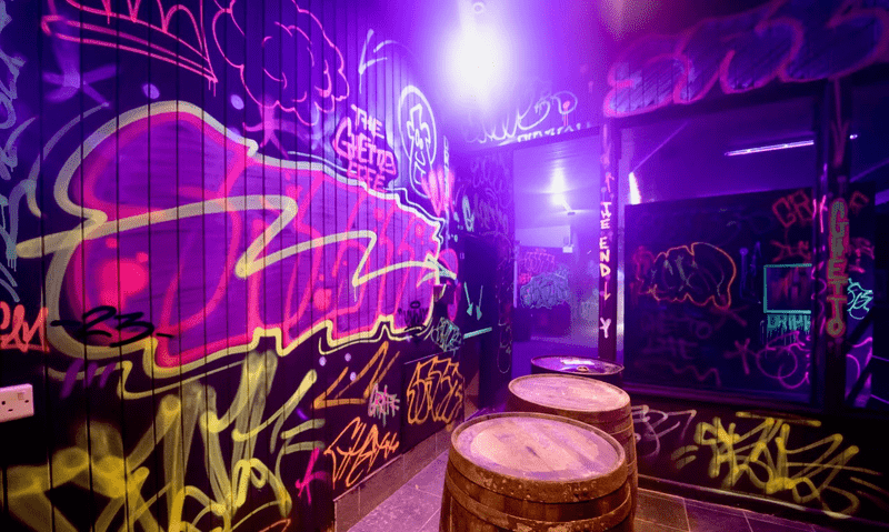 Image of the inside of the laser tag arena with neon lights, barrels and graffiti